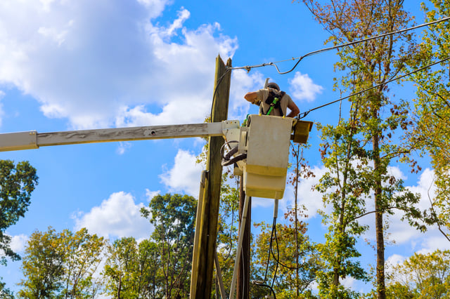 A man is working on a utility pole, standing on a bucket and using a ladder to reach the top. He is wearing a backpack and a helmet for safety. The scene is set against a backdrop of a blue sky.