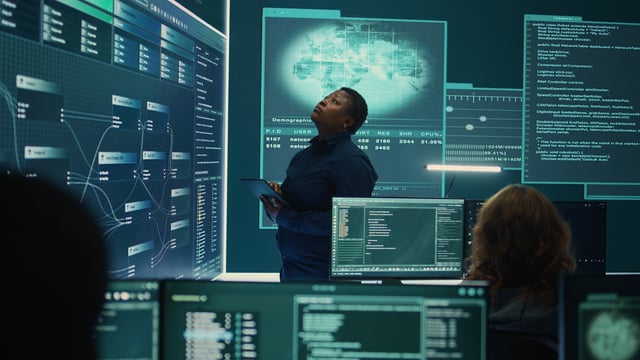 A woman standing in front of a large computer screen, which displays a variety of graphs and data. She appears to be focused on the information displayed on the screen.